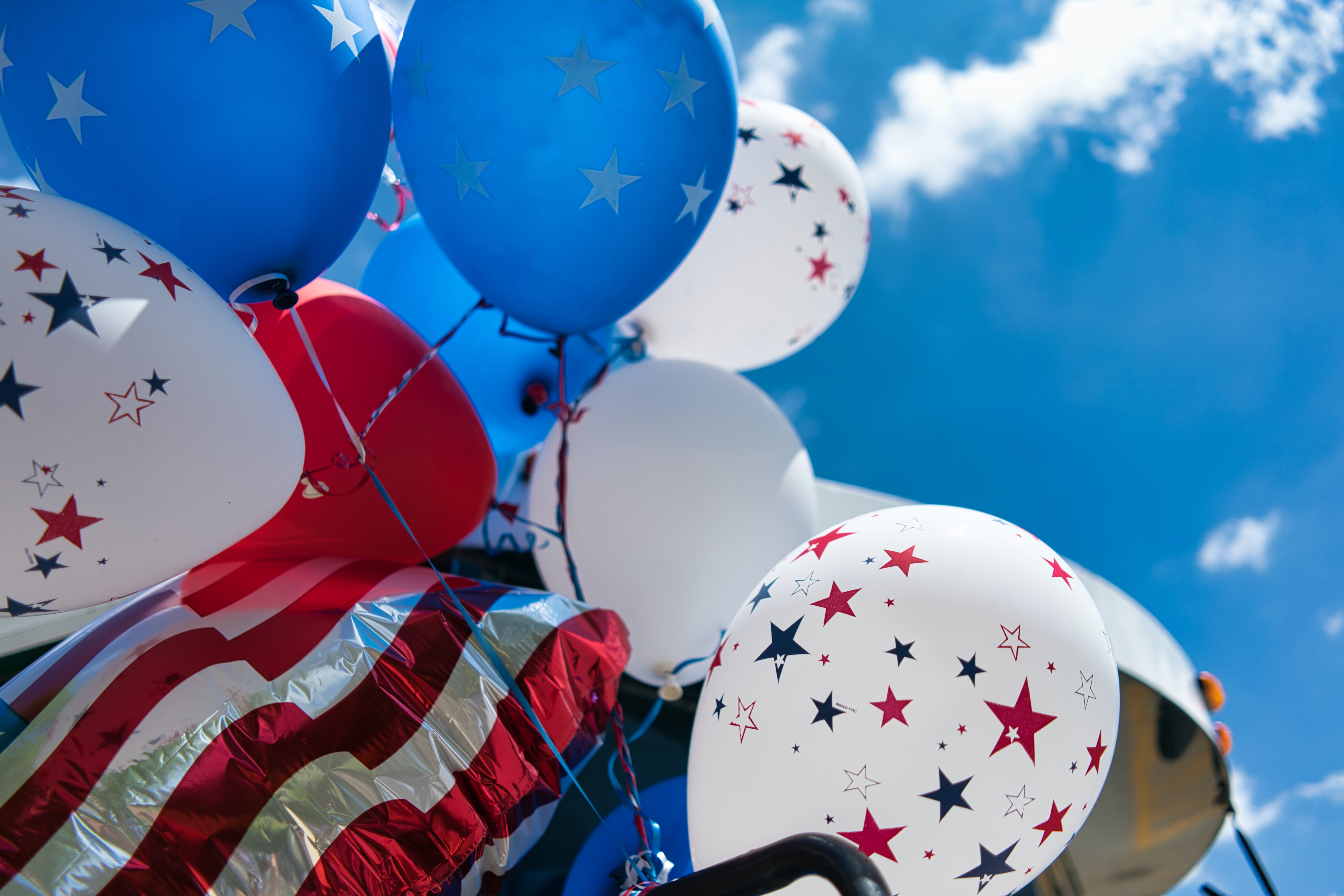 A low-angle close-up view of a festive cluster of red, white, and blue balloons floating against a bright blue sky with soft white clouds. The collection includes white balloons speckled with red and blue stars, solid blue balloons with white stars, and a metallic foil balloon with red and silver stripes.