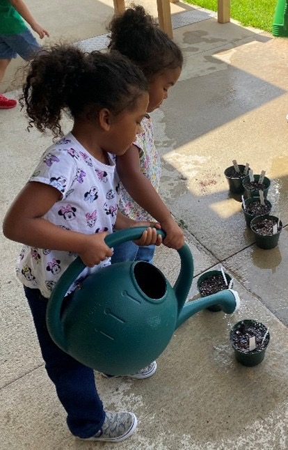 Two young girls standing on a concrete patio under a shaded structure, cooperating to water a line of small potted plant seedlings. One girl in a Mickey Mouse shirt tilts a large green plastic watering can while the other girl guides it.