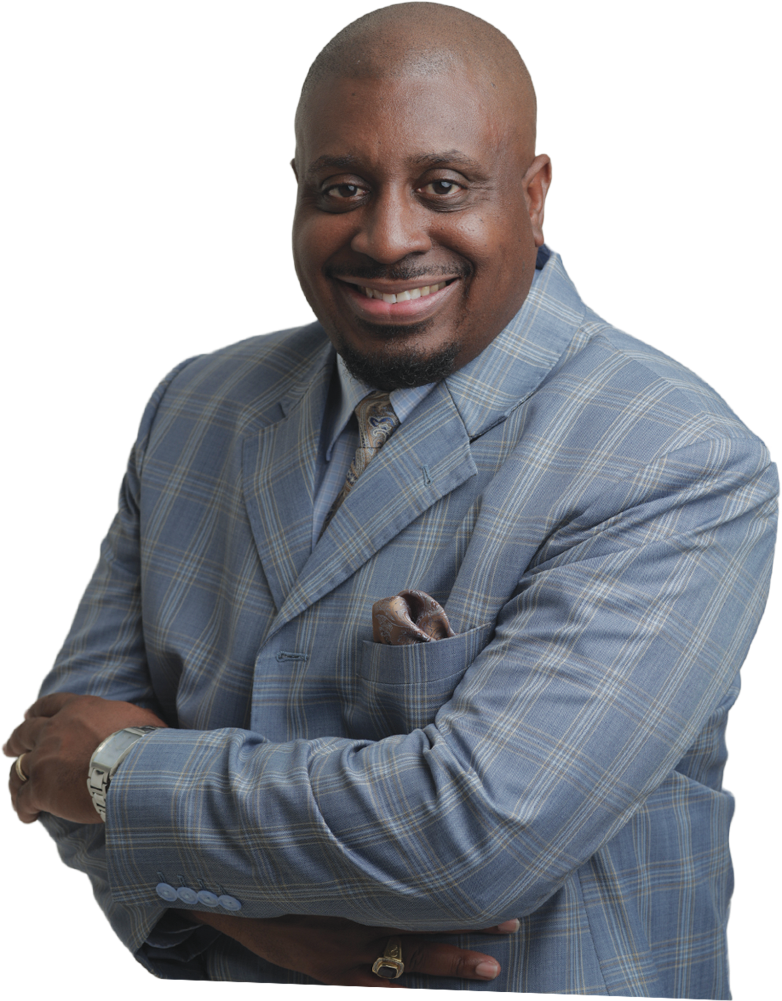 A professional studio portrait of a Black man with a goatee smiling warmly with his arms crossed against a solid black background. He is wearing a light blue and grey plaid suit jacket, a collared shirt, a gold patterned tie, and a matching pocket square.