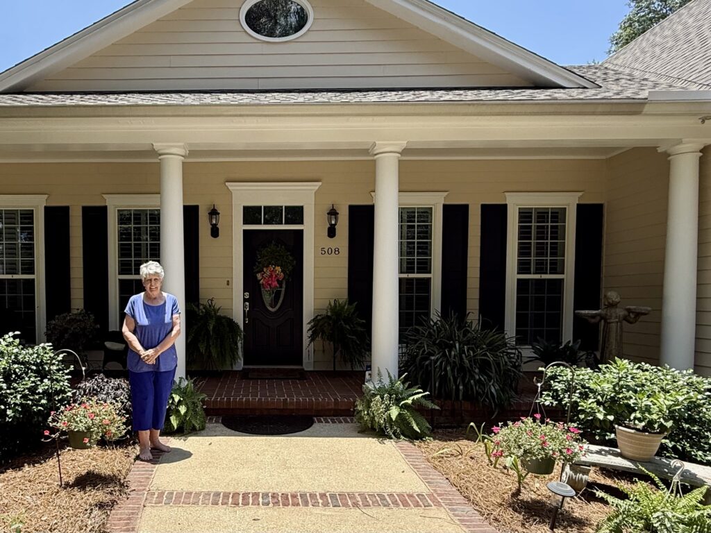 An elderly woman with short white hair standing on the front sidewalk of a beige single-story house with white columns and dark shutters. She is wearing a blue top and blue pants, surrounded by potted plants, hanging flower baskets, and lush green landscaping.