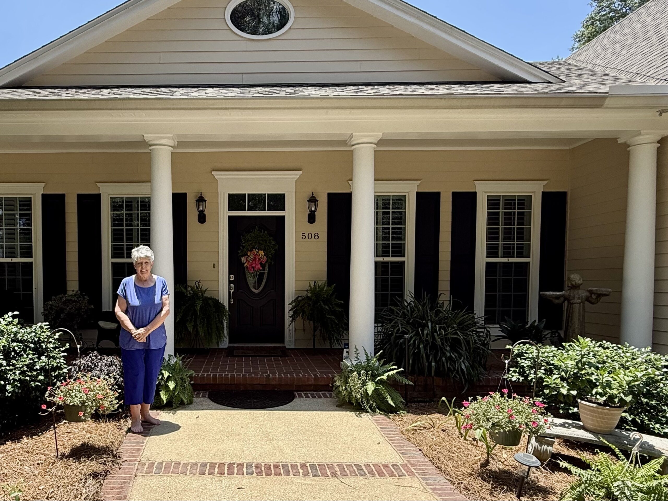 An elderly woman with short white hair standing on the front sidewalk of a beige single-story house with white columns and dark shutters. She is wearing a blue top and blue pants, surrounded by potted plants, hanging flower baskets, and lush green landscaping.