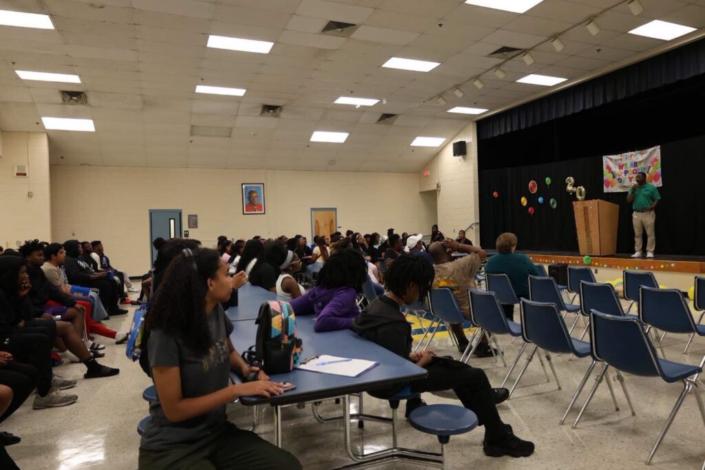 A large group of students seated at cafeteria tables and in rows of chairs in a school auditorium, listening to a man speaking at a wooden podium on a stage. The stage features a black backdrop decorated with colorful paper fans, gold balloons spelling '20', and a partially visible 'We Are Proud of You' banner.