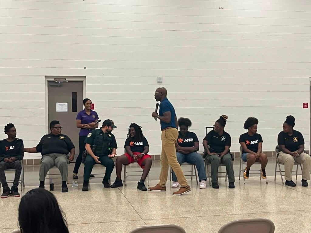 A man standing in front of a group of seated teenagers and police officers in a gymnasium, holding a microphone and addressing the audience.