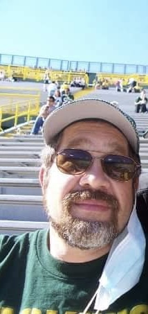 Selfie of a man with a beard, sunglasses, and a baseball cap sitting in stadium bleachers with a face mask tucked around his ear.