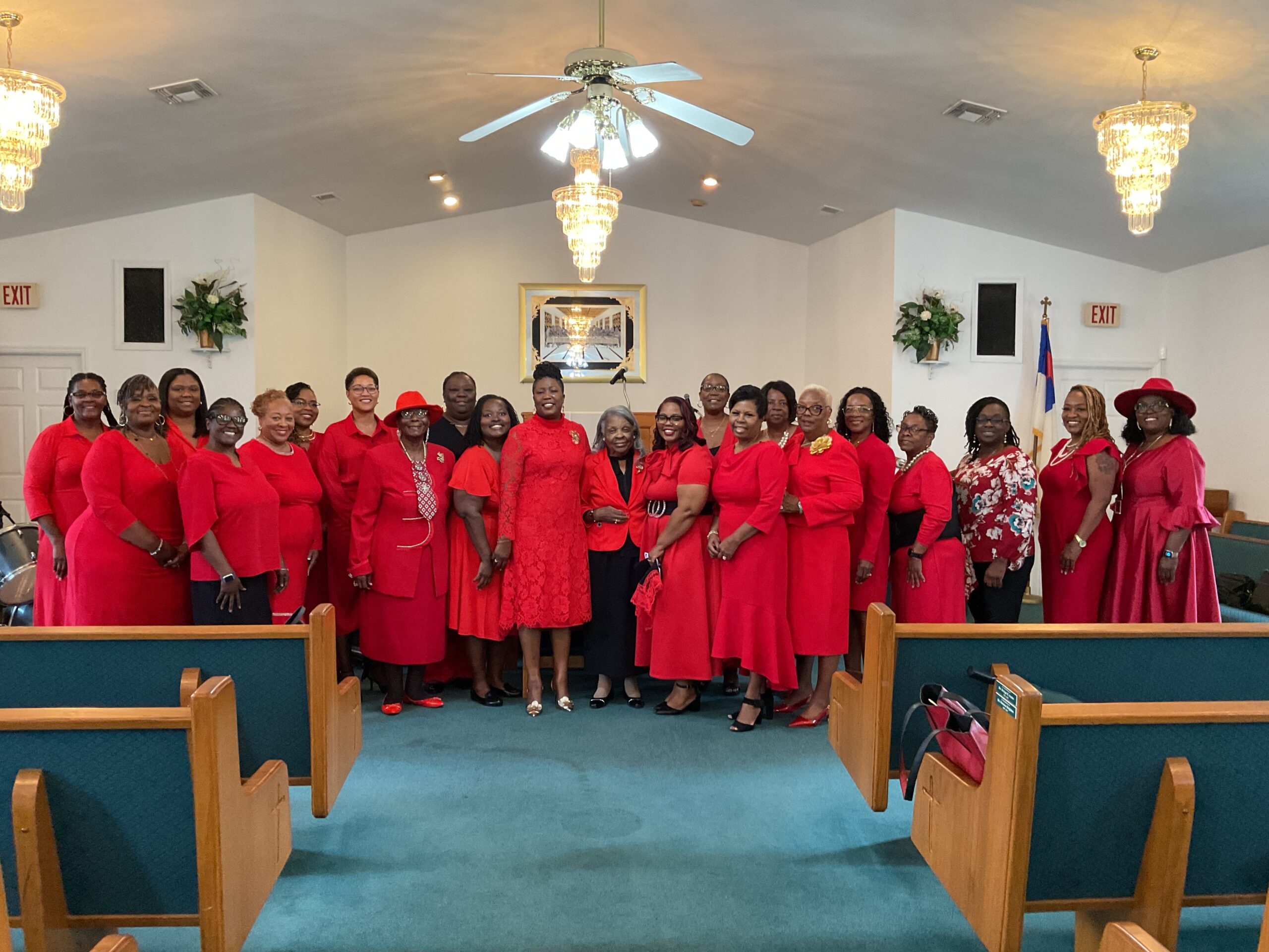 A large congregation group of women wearing matching red dresses standing in rows at the front of a sanctuary between wooden pews under chandelier lights.