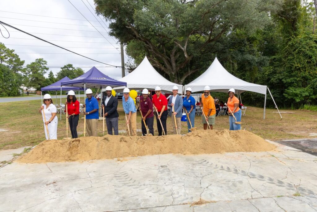 Local officials and project leaders in hard hats breaking ground with shovels at a dirt pile during an outdoor groundbreaking event under canopy tents.