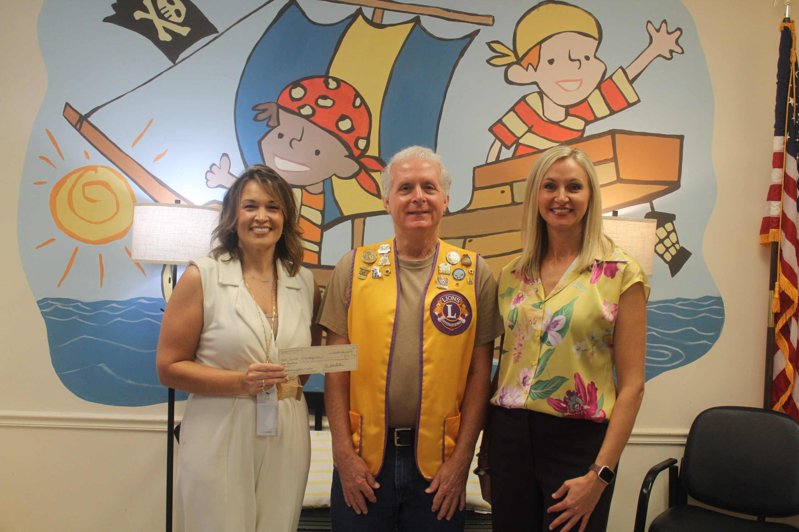 A representative wearing a yellow Lions Club vest presenting a charitable donation check to two women standing in front of a colorful pirate-themed wall mural.