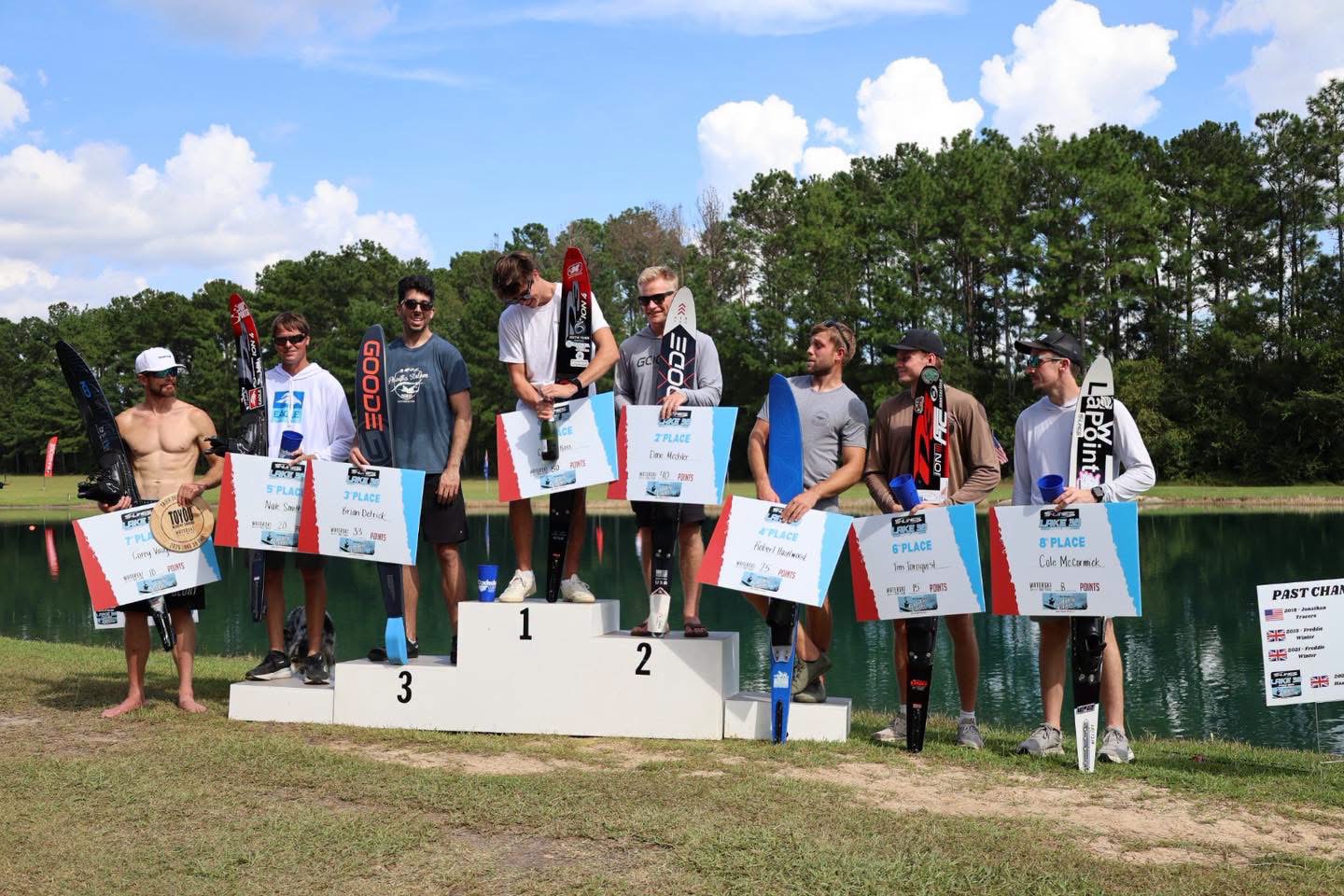 Top eight male water ski competitors standing with their skis and placement signs on a podium in front of a lake under a sunny sky.