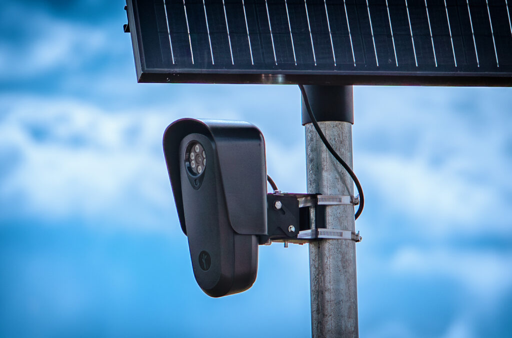 A close-up photograph of a black Flock Safety license plate reader camera and solar panel mounted on a metal pole against a blue sky.