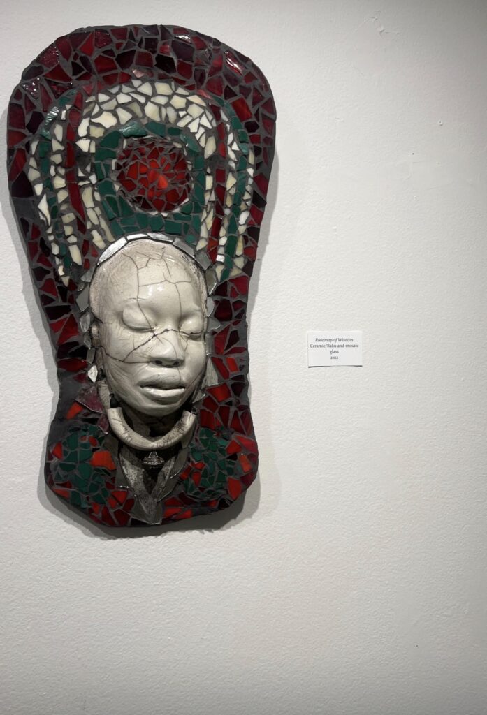 A ceramic and glass mosaic artwork titled 'Roadmap of Wisdom' featuring a white face mask against red and green glass pieces mounted on a wall.