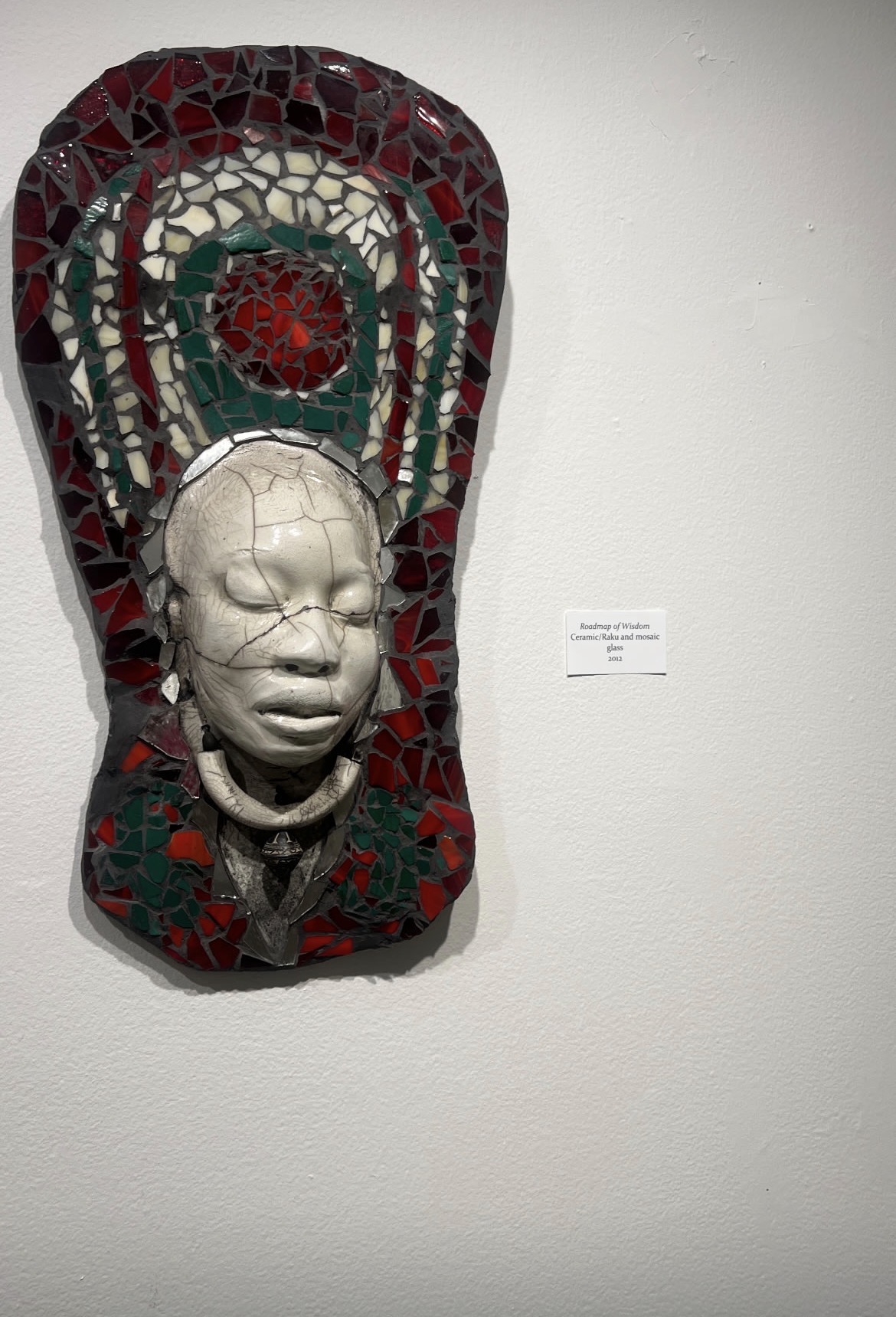 A ceramic and glass mosaic artwork titled 'Roadmap of Wisdom' featuring a white face mask against red and green glass pieces mounted on a wall.