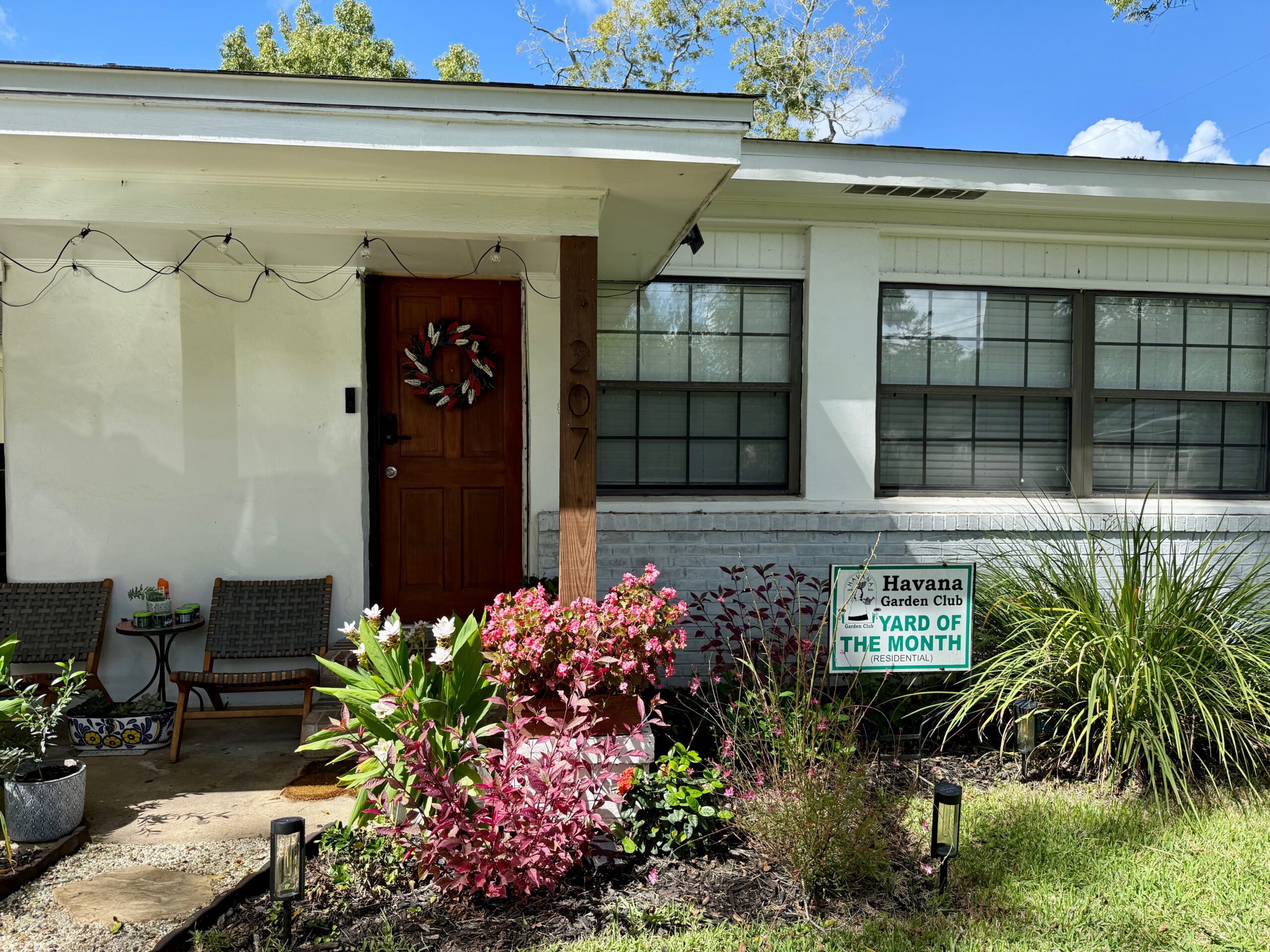 A white house with a dark brown front door decorated with a wreath, featuring a 'Havana Garden Club Yard of the Month' sign surrounded by colorful flowering plants.