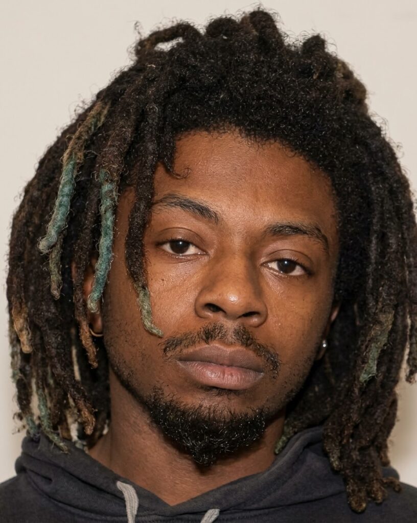 Mugshot photo of a young man with dark dreadlocks and a mustache wearing a black hoodie.