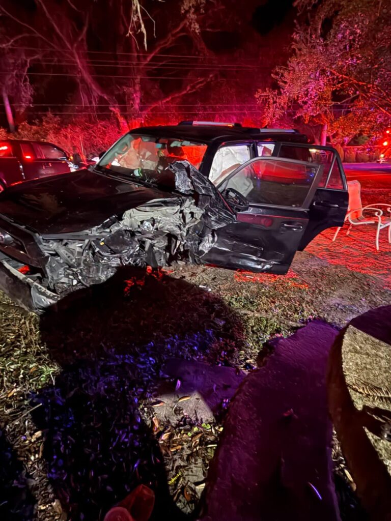 A dark SUV with heavy front-end and side damage from a crash parked at night with emergency lights illuminating the scene.