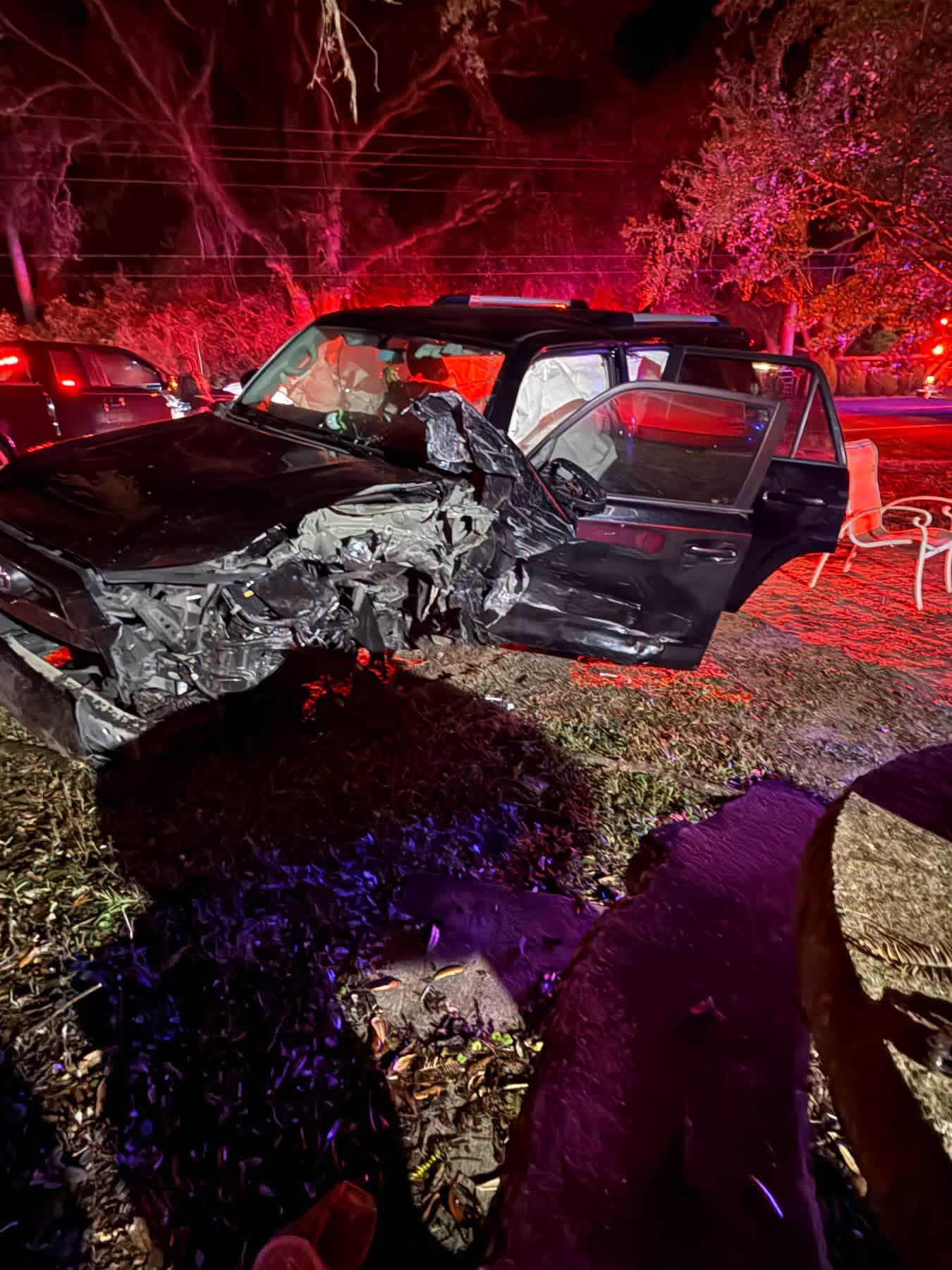 A dark SUV with heavy front-end and side damage from a crash parked at night with emergency lights illuminating the scene.
